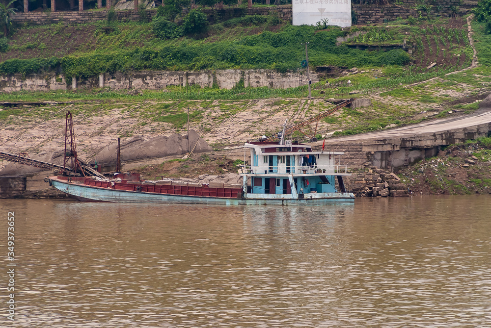 Myaoyinan Chongqing, China - May 8, 2010: Yangtze River. Blue barge docked at shoreline of factory near slipway and transport belt. Brown water up front, green and brown stone slope.