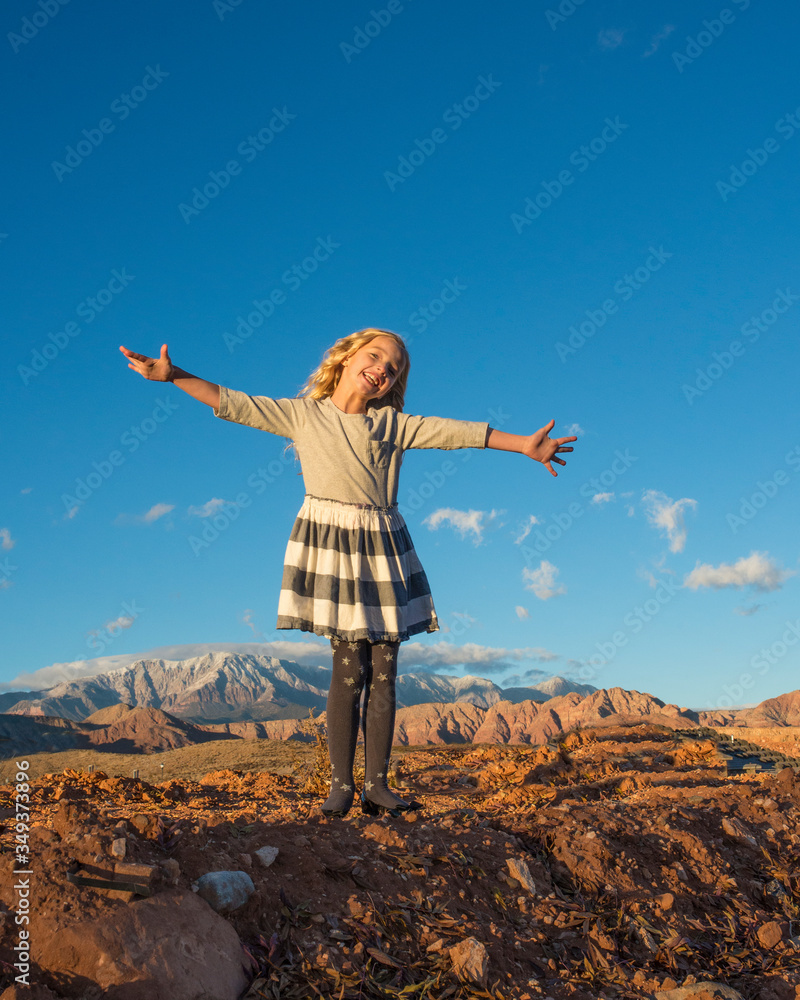 Young girl stands on red rock precipice with arms outstretched in joy. Snow covered mountains and blue sky shown in the background Young girl stands on red rock precipice with arms outstretched in joy. Snow covered mountains and blue sky shown in the background