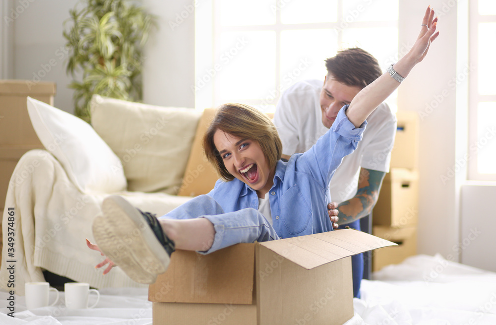 Couple having fun laughing moving into new home, young woman riding sitting in cardboard box while man pushing it Couple having fun laughing moving into new home, young woman riding sitting in cardboard box while man pushing it