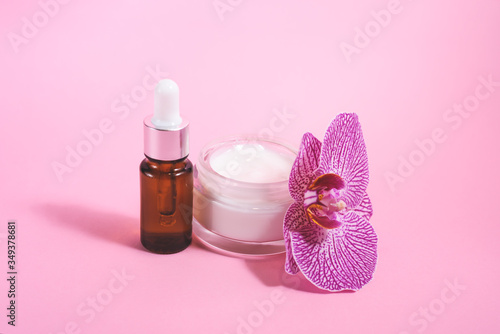 A cosmetic bottle and purple orchid and facial or bode cream on pink background.