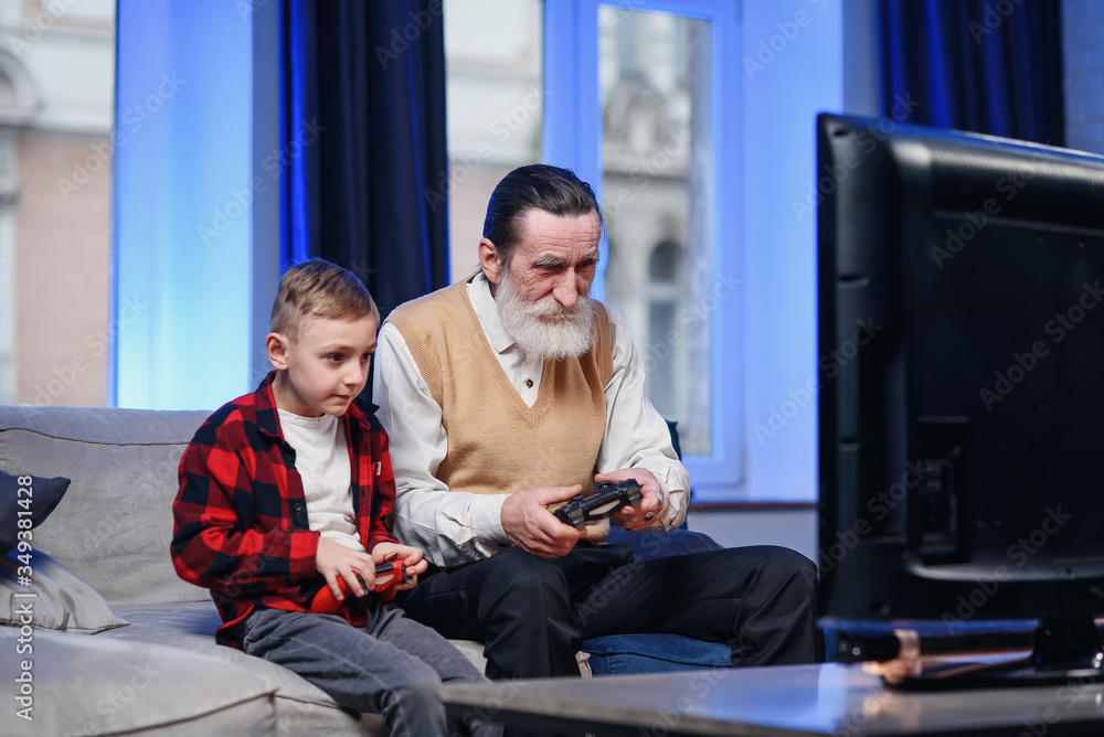 Cute little boy with grandfather sitting on sofa and playing video game with game pad Cute little boy with grandfather sitting on sofa and playing video game with game pad