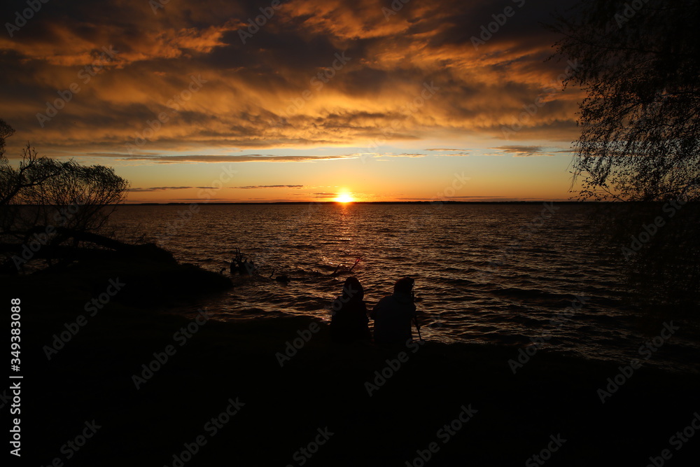 Two people at sunset with a camera sitting on the shore of a raging lake under storm clouds admiring the falling sun from a clear horizon.The fire is burning in half the sky.Russia