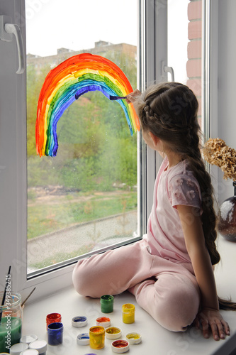 Little smiling girl sits by the window on the windowsill and draws a rainbow on the glass
