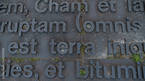 Artistic creation of Latin words in relief, on a metallic structure, covering the ground of a city street, tufts of herbs springing from the work