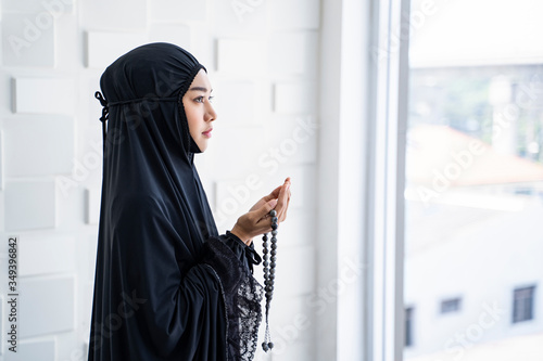 portrait side view of beautiful asian muslim woman looking up palms open to light praying to allah with prayer beads in the hands of the palm, wearing black hijab robe, in prayer room with cool tone