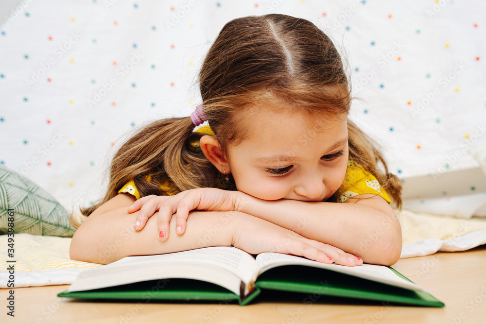 Entertained little girl keen on reading book while lying on the floor. Entertained little girl keen on reading book while lying on the floor.