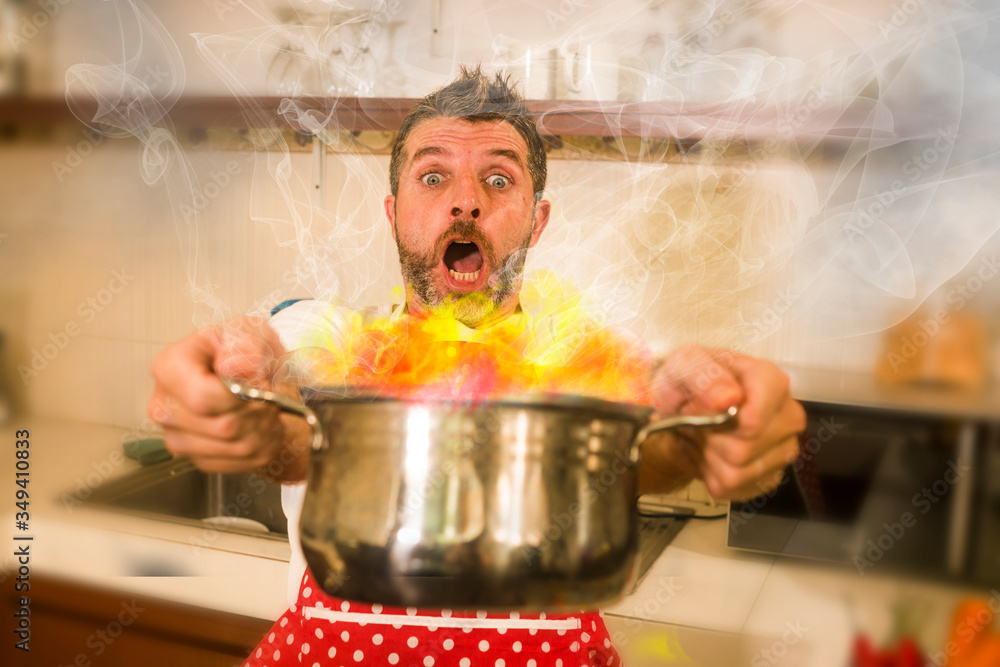 disaster home cook at kitchen- young funny and desperate man in cooking  apron holding pot in flames in stress and fear making a mess of fire and  smoke with food burning Stock