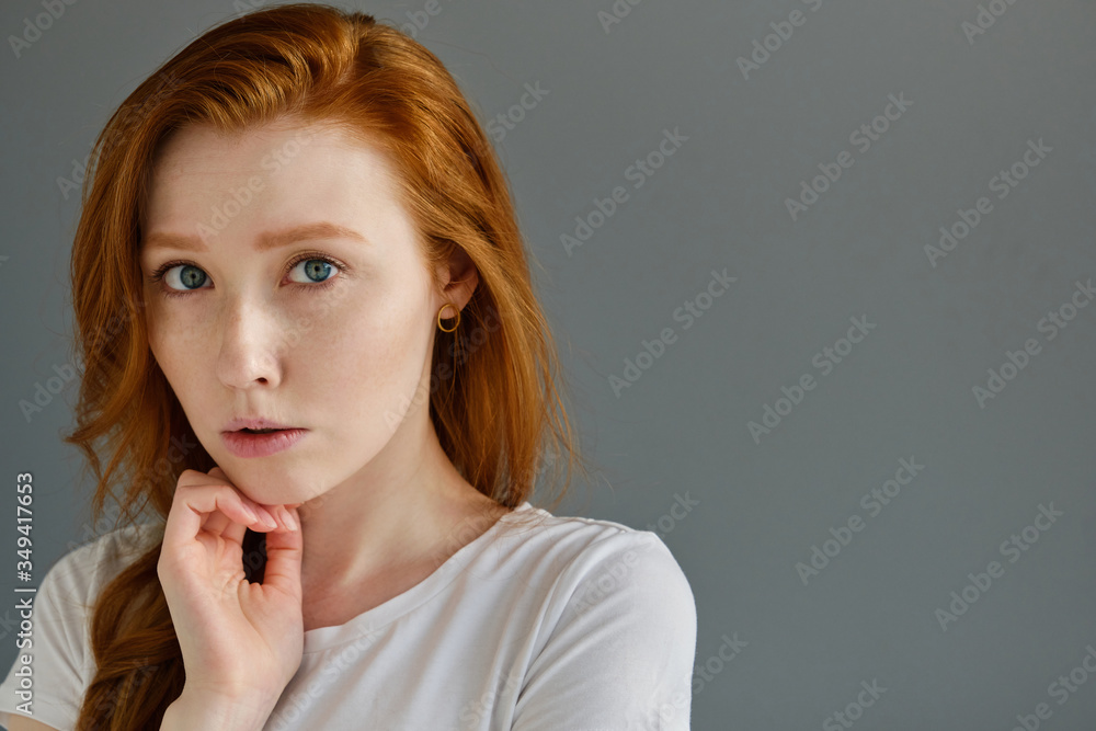 Head shot. A redhead girl with clean skin and blue eyes in a white T-shirt stands on a gray background, backed up chin with hand Head shot. A redhead girl with clean skin and blue eyes in a white T-shirt stands on a gray background, backed up chin with hand