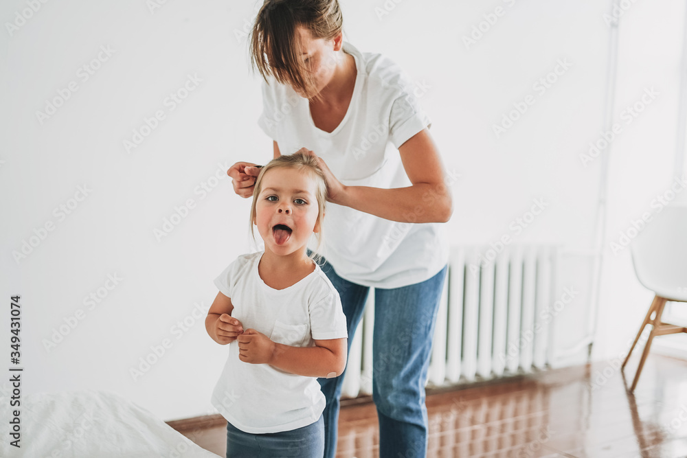 Fototapeta premium Funny moment of real life when young mother doing to her daughter toddler girl pony tail in the bright room