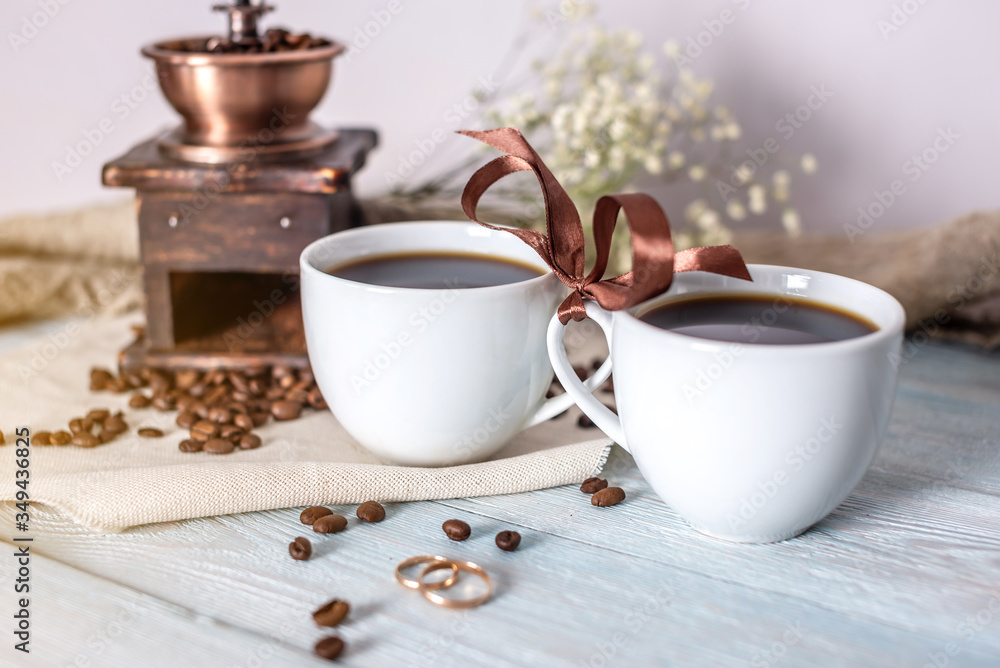 Couple of cups of fresh, aromatic black coffee are tied together with a ribbon. Wedding rings are placed in front of the mugs Couple of cups of fresh, aromatic black coffee are tied together with a ribbon. Wedding rings are placed in front of the mugs