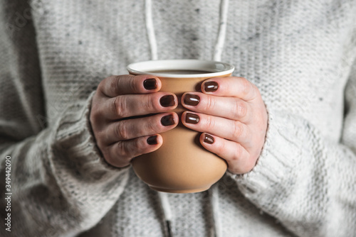 A woman wearing in a light cozy sweater is holding a cup of aromatic black coffee. The nails are coated with brown nail polish. Concept of coffee manicure.