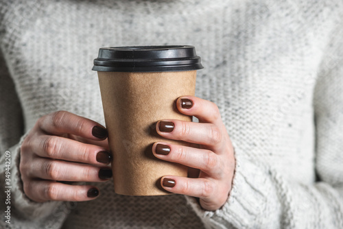 A woman wearing in a light cozy sweater is holding a paper cup of aromatic coffee. The nails are coated with brown nail polish. Concept of elegance and stylish coffee manicure. photo