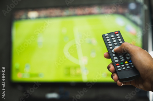 Young man holding television remote control watching football program. Hands pointing to tv screen set and turning it on or off select channel watching tv in the living room relax.