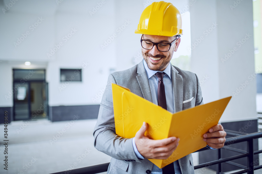 Attractive caucasian elegant architect in suit and with helmet on head leaning on railing and looking at documents in folder. Attractive caucasian elegant architect in suit and with helmet on head leaning on railing and looking at documents in folder.