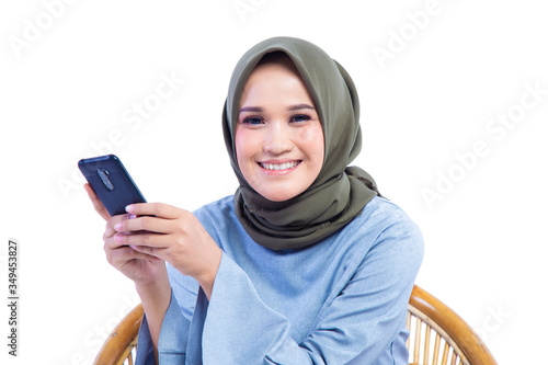Beautiful Hijab woman sitting in a chair while holding a smart phone with smile expression, isolated on white background.