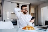 Young caucasian serious businessman with eyeglasses sitting at table in dining room and reading news on smart phone. On table are breakfast and laptop.