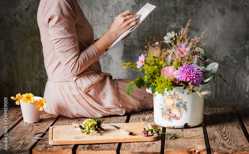 female artist draws a composition of wild flowers in a pot in a rustic style, a sketchbook on a table with pencils.
