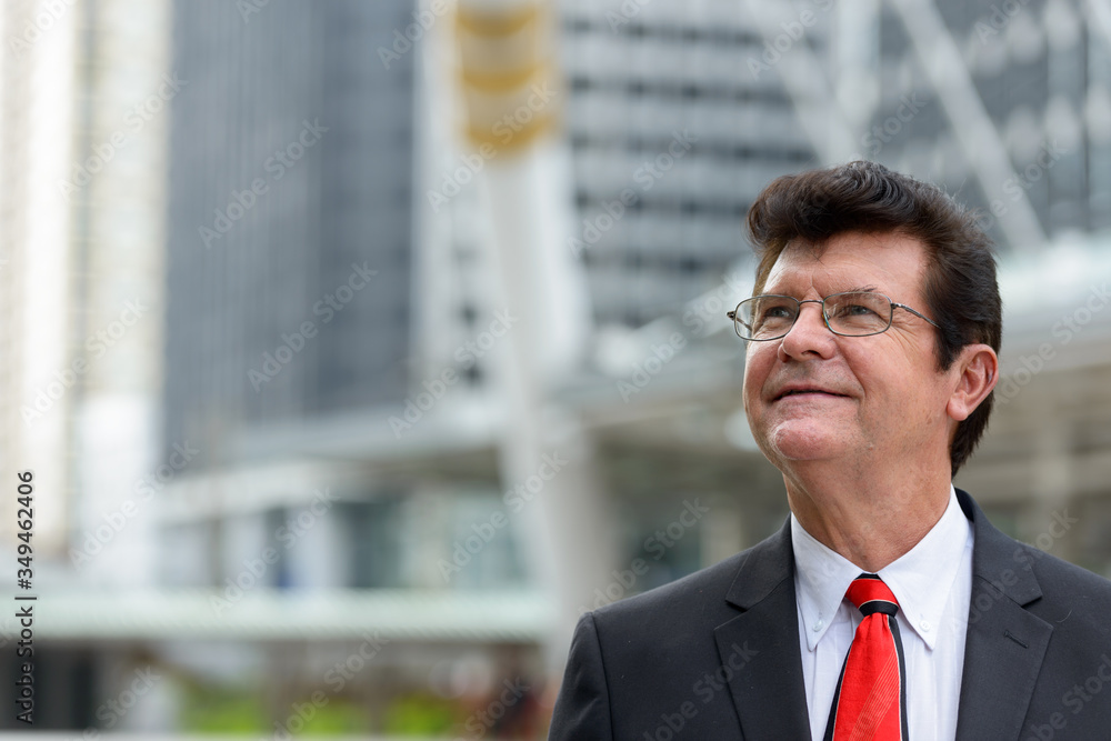 Mature happy businessman thinking in front of modern building at Bangkok city