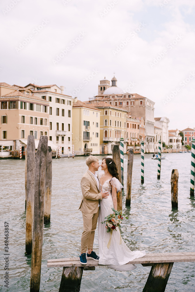 Italy wedding in Venice. The bride and groom are standing on a wooden pier for boats and gondolas, near the Striped green and white mooring poles, against backdrop of facades of Grand Canal buildings.