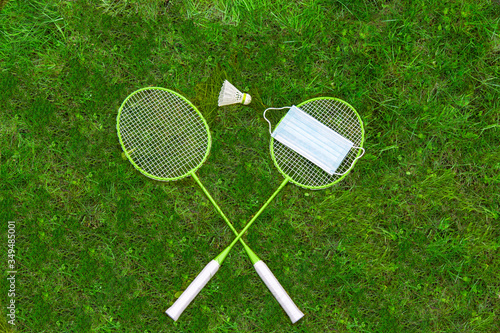 Coronavirus protection sing. Badminton rackets and shuttlecock on green grass. Medical mask on one racket. view from above. ban