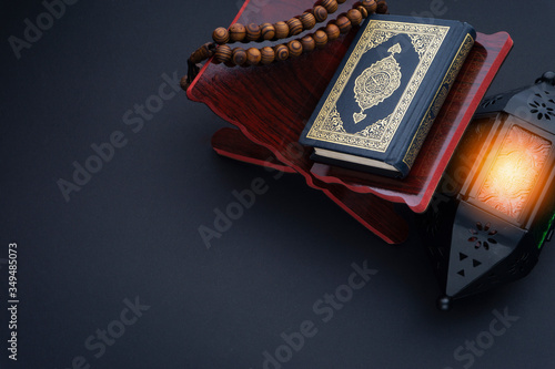 Holy Al Quran with written arabic calligraphy meaning of Al Quran, lantern lamp and rosary beads or tasbih on black background.