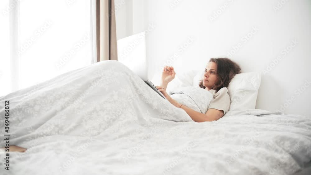A young sleepy woman in pajamas working at home using her laptop while sitting on her bed in a bedroom with a cup of morning coffee. A young sleepy woman in pajamas working at home using her laptop while sitting on her bed in a bedroom with a cup of morning coffee.
