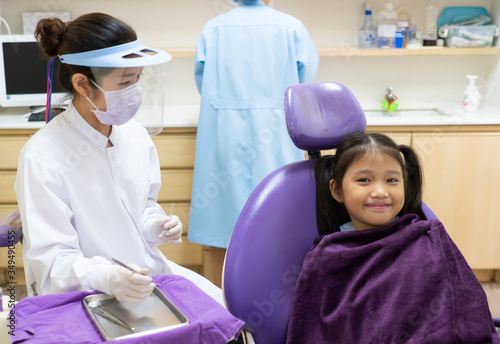 dentist using dental tools to clean teeth of asian child and treat tooth decay in the clinic with asisstance standing behind the patient. dentistry and healthcare concept