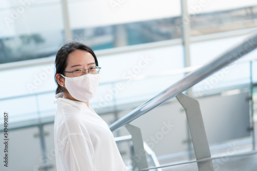 young asian woman with smiling face and wearing face mask as a social distancing guideline. She is using stairway to go upstair to the sky train station. new normal, covid-19, coronavirus concept