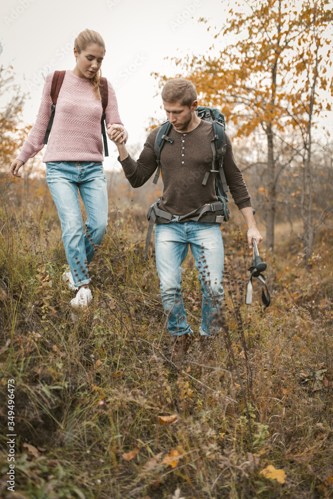 Man is supporting a beautiful blonde with a backpack walking together in the wild nature in autumn