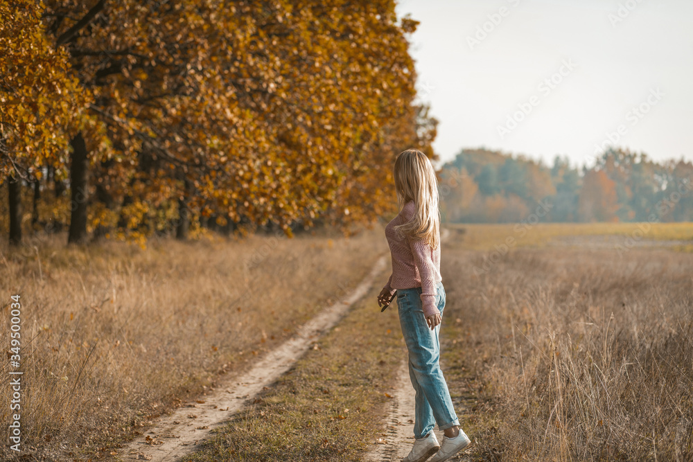 Rear view of young woman in casual clothes tiptoeing in nature, charming young blonde walking along a dirt road near the autumn forest Rear view of young woman in casual clothes tiptoeing in nature, charming young blonde walking along a dirt road near the autumn forest