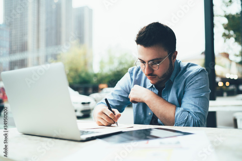 Serious man writing documents sitting at table with laptop in outside cafe