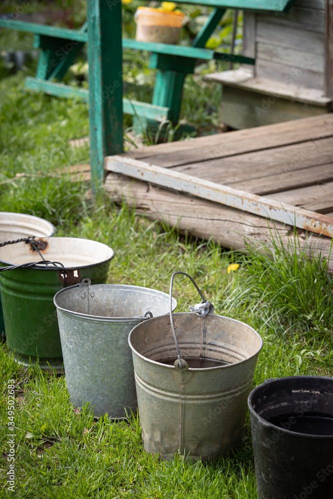 Many different buckets stand in a row to collect rainwater. Buckets of water stand on the grass in the garden next to the house. Village concept Many different buckets stand in a row to collect rainwater. Buckets of water stand on the grass in the garden next to the house. Village concept