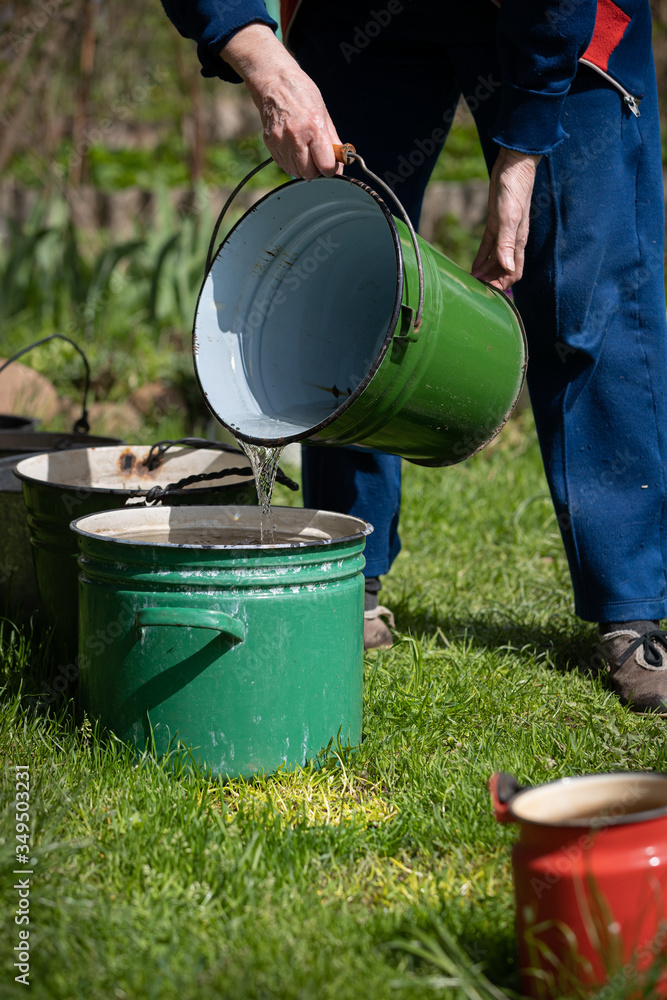 Several old buckets are standing on the grass in sunny weather. Grandmother pours the collected water into an empty bucket. Collecting rainwater in buckets in the village. Buckets are in one line. Several old buckets are standing on the grass in sunny weather. Grandmother pours the collected water into an empty bucket. Collecting rainwater in buckets in the village. Buckets are in one line.
