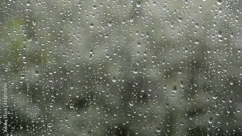 strong rain water drops fall, hit and flow on glass window surface, bad windy summer day weather and green trees blurred background, selective focus of closeup full hd stock video footage in real time
