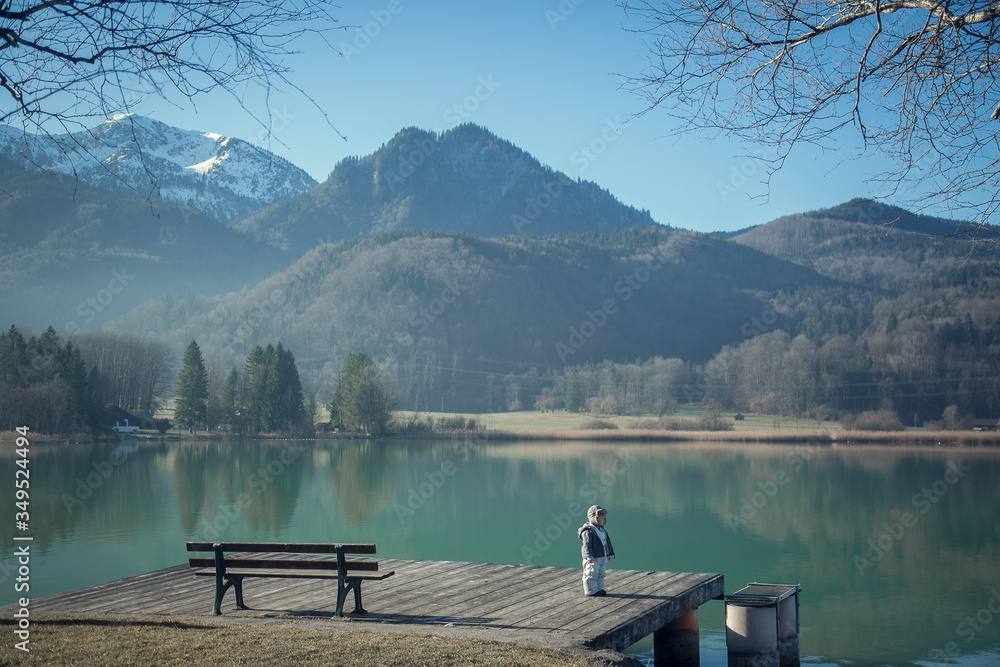 A child stands on a bridge near a bench by a mountain lake in spring