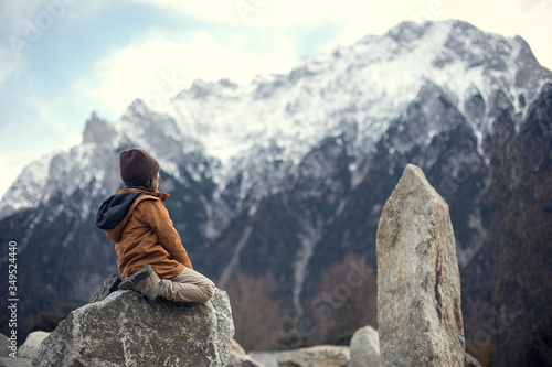 Photography A boy in a hat and a jacket sits on a stone in the mountains strewn with snow an