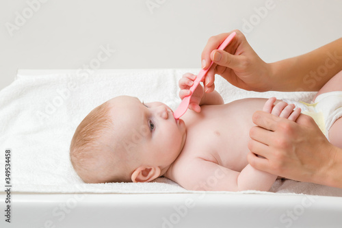 Young mother hands holding pink plastic spoon with syrup. Infant lying down on changing table and receiving vitamins. Side view. Closeup.