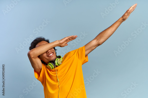 Cheerful African-American young guy joyfully raises hands up covering face as dub dancing sign posing on blue background with headphones in yellow t-shirt. Сoncept of music subscription, online radio photo