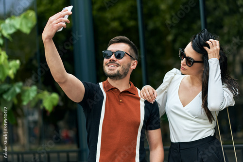 Beautiful young woman and handsome man standing outdoor on the city street with smartphone for taking selfie. Young couple walks at the city street, making self portrait on mobile phone.