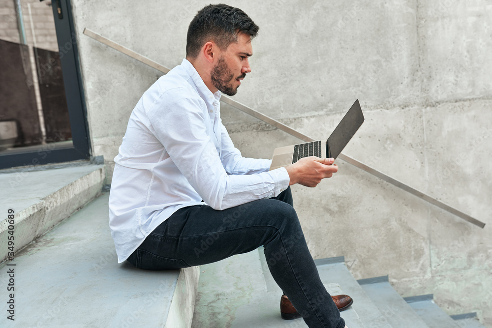Side view shot of young bearded man sitting on the concrete stairs in urban city center outdoor working on the laptop. Hipster male using laptop for chatting online with friends, browsing on Internet Side view shot of young bearded man sitting on the concrete stairs in urban city center outdoor working on the laptop. Hipster male using laptop for chatting online with friends, browsing on Internet
