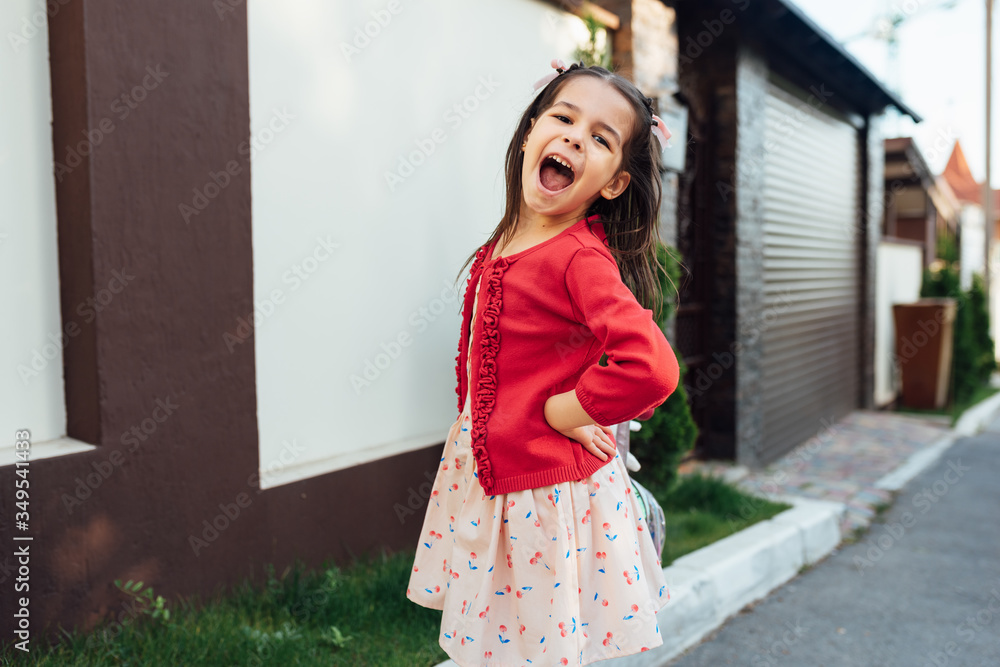Image of cute little girl laughing during coming home after school day stading near her home outside. Smiling kid playing outdoor, has joyful expression. Happy child smiling broadly and posing outdoor Image of cute little girl laughing during coming home after school day stading near her home outside. Smiling kid playing outdoor, has joyful expression. Happy child smiling broadly and posing outdoor