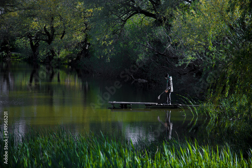 A boy with a bucket and a net on an old wooden pier on a beautiful green lake surrounded by trees