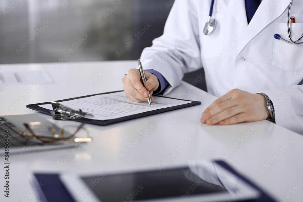 Unknown male doctor sitting and working with clipboard of medication history record in clinic at his working place, close-up. Young physician at work. Perfect medical service, medicine concept Unknown male doctor sitting and working with clipboard of medication history record in clinic at his working place, close-up. Young physician at work. Perfect medical service, medicine concept