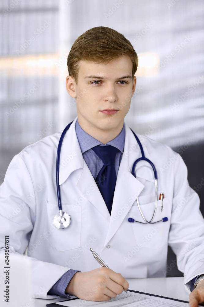 Blond male doctor sitting and working with clipboard in clinic at his working place, close-up. Young physician at work. Perfect medical service, medicine concept Blond male doctor sitting and working with clipboard in clinic at his working place, close-up. Young physician at work. Perfect medical service, medicine concept