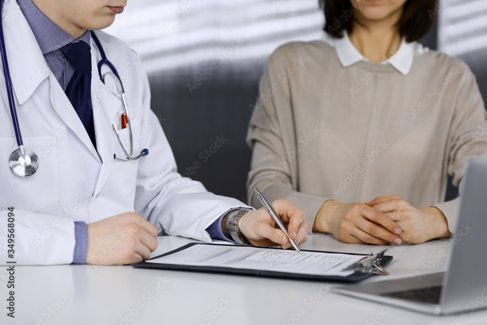 Obraz premium Unknown male doctor and patient woman discussing something while sitting in clinic and using clipboard. Best medical service in hospital, medicine, pandemic stop