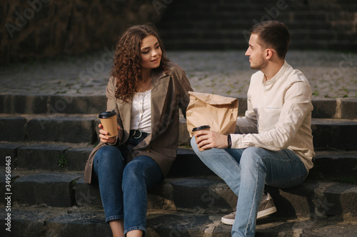 Young couple takaaway food and sit outdoors on stairs in city. Stylish couple drink coffe and wait friends