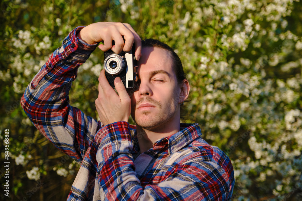 Hipster in flannel shirt is photographing flowering trees with his film soviet camera in May.
