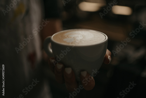 A female barista holds a cup of hot latte in her hand in a cafe. A waitress preparing a customer order in the evening.