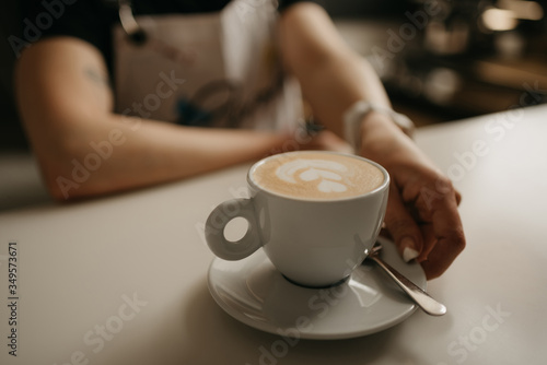 A female barista holds out a cup of hot latte with a saucer and spoon to a client in a cafe. A barista preparing a customer order.