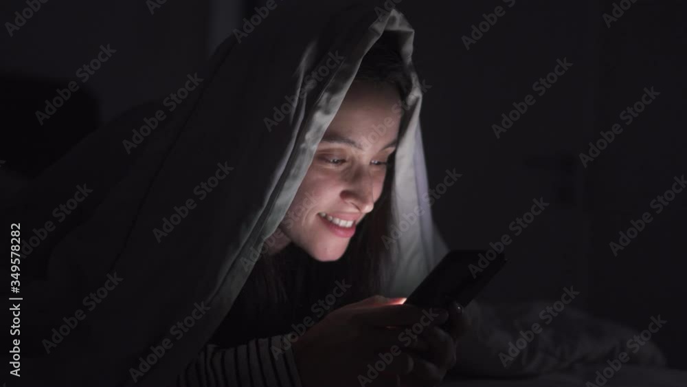 Woman is browsing social media in mobile phone at night under the blanket, Face close-up.
Woman is browsing social media in mobile phone at night under the blanket, Face close-up.